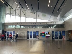 ردهة مركز أريزونا للعلوم ذات النوافذ الكبيرة والجدران الخرسانية والعروض على طول الجدار الخلفي؛ مجموعة من الزوار تقف على اليسار بالقرب من أحد المعروضات